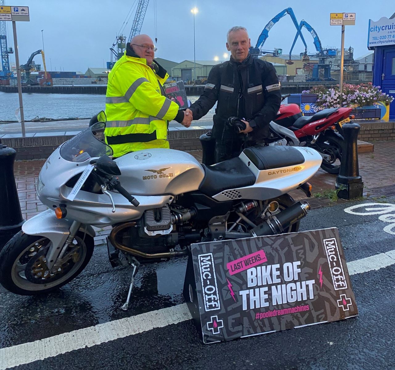 Two men stood next to bike at Poole quay on a rainy evening 